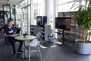 Heller Wartebereich im Mercedes-Autohaus mit Glasfront. Ein junger Mann sitzt an einem runden Tisch und liest eine Broschüre. Neben ihm stehen weitere Stühle, ein kleiner Tisch mit einer Pflanze, ein Kaffeeautomat mit Tassenregal und ein großer Fernseher. Rechts im Bild eine große grüne Pflanze in einem grauen Topf.