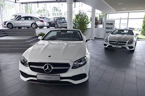 Heller, moderner Mercedes-Benz-Showroom mit großen Glasfronten. Im Vordergrund ein weißes E-Klasse Cabriolet, dahinter ein silbernes Cabriolet. Auf einer erhöhten Fläche stehen weitere Mercedes-Fahrzeuge. Viel Tageslicht, freundliche und offene Atmosphäre.