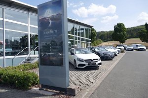 Reihe von Mercedes-Benz Gebrauchtwagen (Junge Sterne) auf dem Parkplatz eines Autohauses. Im Vordergrund steht ein großes Schild mit dem Logo und der Aufschrift ‚Junge Sterne – Die besten Gebrauchten von Mercedes-Benz‘. Im Hintergrund moderne Glasfassade des Autohauses, Bäume und Felder bei sonnigem Wetter.