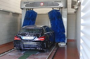 Ein schwarzer Mercedes-Benz wird in einer automatischen Waschanlage mit blauen Bürsten gereinigt. Das Fahrzeug ist mit Schaum bedeckt, die Anlage befindet sich in einer gefliesten Halle.