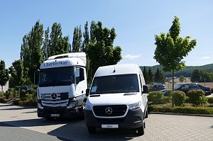 Zwei weiße Mercedes-Benz Nutzfahrzeuge stehen nebeneinander auf einem Parkplatz. Links ein großer CharterWay-Lkw, rechts ein Transporter. Im Hintergrund grüne Bäume, Autos und leicht hügelige Landschaft unter blauem Himmel.