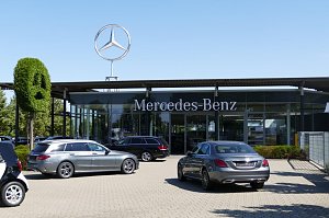 Außenansicht eines Mercedes-Benz-Autohauses mit großem Schriftzug ‚Mercedes-Benz‘ an der Glasfassade. Darüber ein großes silbernes Mercedes-Logo auf dem Dach. Vor dem Gebäude stehen mehrere silberne und schwarze Mercedes-Fahrzeuge. Links eine grüne, in Form des Markenlogos geschnittene Buchsbaumfigur.