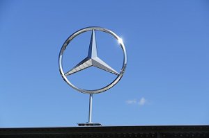 Großes silbernes Mercedes-Benz-Logo in Form des dreizackigen Sterns, montiert über einem Gebäudedach. Strahlend blauer Himmel im Hintergrund, die Sonne spiegelt sich am Rand des Logos.