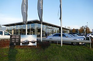Mercedes-Benz Autohaus mit großer Glasfassade und davor geparkten Fahrzeugen, vor allem silberfarbene Limousinen. Im Vordergrund ein Schild mit der Aufschrift ‚Junge Sterne glänzen länger‘ und einem Bild von Mercedes-Fahrzeugen. Drei hohe Fahnenmasten mit Mercedes-Benz-Fahnen. Grünfläche mit Gras und Sträuchern, blauer Himmel.