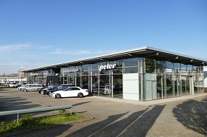Autohaus Peter mit moderner Glasfassade und der Aufschrift ‚Gebrauchtwagen‘. Vor dem Gebäude sind mehrere Mercedes-Fahrzeuge in verschiedenen Farben geparkt, darunter ein weißer Wagen im Vordergrund. Weitläufiger Parkplatz, blauer Himmel ohne Wolken.