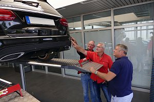 Drei Männer stehen in einem hellen Werkstattbereich unter dem Heck eines schwarzen Mercedes-AMG, der auf einer Hebebühne angehoben ist. Einer der Männer zeigt auf eine Stelle am Fahrzeug, die anderen beiden schauen interessiert zu.