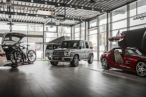 Innenraum einer modernen Mercedes-Benz-Ausstellungshalle mit großen Glasfronten. Zu sehen sind ein weißer Kombi mit offenem Kofferraum und Fahrradträger, ein silberner Mercedes G-Klasse SUV und ein roter Sportwagen mit geöffneten Flügeltüren. Heller, industrieller Boden und Decke mit Lampen.