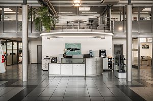 Moderner Empfangsbereich eines Autohauses mit halbkreisförmigem Tresen, Druckern, Kaffeemaschine und Wasserspender. An der Wand hängt ein Bild mit Mercedes-Autos. Darüber eine Galerie mit Geländer und Tisch mit Stühlen. Der Raum hat große Fenster, graue Fliesen und eine hohe Decke.