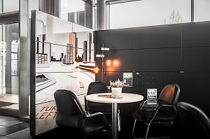 Gemütliche Sitzecke mit rundem Tisch und vier schwarzen Lederstühlen in einem Autohaus. Auf dem Tisch steht eine kleine Pflanze. Im Hintergrund eine Wand mit großem Stadtmotiv in Weiß- und Orangetönen. Helle Fensterfront und moderner schwarzer Wandbereich.