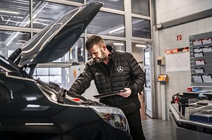 Werkstattmitarbeiter im Mercedes-Benz Autohaus prüft den Motorraum eines Fahrzeugs mit geöffnetem Vorderhaube. Er trägt eine schwarze Jacke mit Mercedes-Logo und hält ein Tablet. Im Hintergrund moderne Werkstatteinrichtung mit Dokumentenfächern, Kalender und Glastüren.