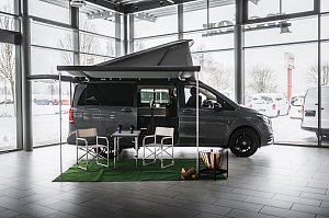 Grauer Mercedes-Benz Camper-Van mit ausgefahrener Markise und aufgeklapptem Schlafdach in einer hellen Ausstellungshalle. Vor dem Van stehen zwei Campingstühle und ein Tisch auf grünem Teppich, daneben ein dekorativer Korb mit Holzscheiten. Draußen ist Schnee zu sehen.