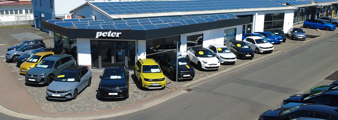 Au&szlig;enansicht des Opel-Autohauses in Witzenhausen mit mehreren bunten Fahrzeugen (u.a. rot, orange, blau, silber) auf dem Parkplatz vor dem Geb&auml;ude. &Uuml;ber dem Eingang ist ein gro&szlig;es gelbes Opel-Logo zu sehen. Der Himmel ist blau und die Sonne scheint.