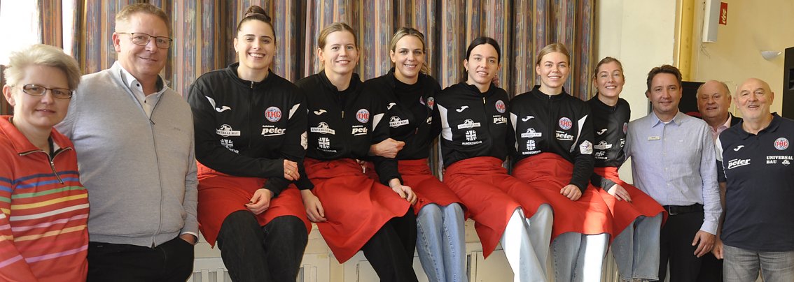 Gruppenfoto im Restaurant der Herzen: 6 junge Frauen mit schwarzen Sportoberteilen und roten Sch&uuml;rzen sitzen nebeneinander auf einer Heizung. Links und rechts stehen jeweils mehrere Personen. Alle blicken in die Kamera und l&auml;cheln.