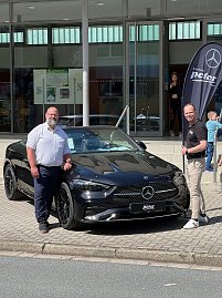 Zwei Männer stehen vor einem schwarzen Mercedes-Benz Coupé und lächeln in die Kamera. Rechts im Hintergrund ist eine Mercedes-Fahne, dahinter Glasfassade der Stadthalle. (Foto: Autohaus Peter)