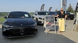 „Eine Frau mit schwarzem Polohemd, gelber Hose und Sonnenbrille steht neben einem Aufsteller mit Werbung für den neuen Mercedes CLA. Links im Bild ein schwarzer Mercedes CLA, daneben weitere Mercedes-Modelle in Weiß und Schwarz. Im Hintergrund wehen schwarze Fahnen mit Mercedes-Logo. Aufnahme beim Leichtathletikmeeting in Sondershausen. (Foto: Autohaus Peter)