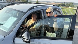 Ein Interessent sitzt am Steuer eines silbernen Mercedes GLB 220 4MATIC, während ihn eine Peter-Mitarbeiterin mit Sonnenbrille und schwarzem Polohemd freundlich berät. Auf der Windschutzscheibe ist ein Datenblatt mit Fahrzeugdetails sichtbar. Im Hintergrund ein Stadion mit Tribüne und grüner Rasen.. (Foto: Autohaus Peter)