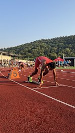 „Sprinter im roten Trikot steht im Startblock auf Bahn 5, bereit für den Lauf. Weitere Athleten bereiten sich auf den benachbarten Bahnen vor. Zuschauer und Kampfrichter am Rand der Tartanbahn, im Hintergrund grüne Hügel. (Foto: Autohaus Peter)