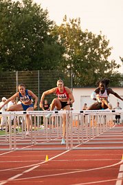 Drei Athletinnen im Hürdenlauf springen gleichzeitig über eine Hürde auf der roten Tartanbahn. Im Vordergrund sind die Hürden mit der Aufschrift Sportzentrum am Göldner zu sehen. Zuschauer und Kampfrichter im Hintergrund. (Foto: Autohaus Peter)