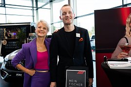 Eine Frau (Moderatorin Mandy Kossira) in lila Blazer und pinkem Top steht neben einem Mann im schwarzen Anzug. Beide lächeln. Neben ihnen ein Aufsteller mit der Aufschrift „Mein Fischer“. (Foto: Buchardt | Autohaus Peter )