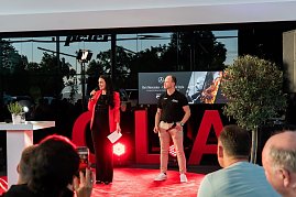 Christina Dost im roten Blazer spricht mit einem Mikrofon auf der Bühne. Neben ihr steht ein Mann im schwarzen Polohemd mit Autohaus-Logo. Hinter ihnen eine große LED-Wand mit „Hes Mercedes  Cars, BBQ & Style“. (Foto: Buchardt | Autohaus Peter )