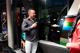 Ein Mann in schwarzem Hemd erklärt mit Mikrofon verschiedene Lack- und Folienmuster an einer Autotür. Daneben bunte Farbfolienstreifen. (Foto: Buchardt | Autohaus Peter )