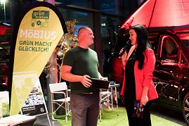 Christina Dost im roten Blazer moderiert neben einem Mann im grünen T-Shirt vor einem gelben Banner mit der Aufschrift „Möbius  Grün macht glücklich!“. Hinter ihnen ein schwarzer Mercedes-Camper. (Foto: Buchardt | Autohaus Peter )