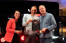 Christina Dost im roten Blazer steht mit Andreas Peter und einem Gewinner auf der Bühne. Gemeinsam halten sie einen großen Gutschein mit Nürburgring-Aufdruck in die Kamera. (Foto: Buchardt | Autohaus Peter )