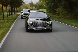 Ein schwarzer Mercedes-Maybach S-Class fährt auf einer Landstraße, gefolgt von einem Mercedes-Maybach SUV. Beide Fahrzeuge sind von vorne zu sehen, mit dem S-Class im Vordergrund und dem SUV dahinter. Die Straße wird von Bäumen gesäumt, und die Farben des Herbstes sind im Hintergrund sichtbar.