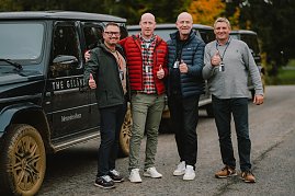 Vier Männer stehen lächelnd vor einem schwarzen Mercedes-Benz G-Class mit schlammigen Reifen. Alle tragen Freizeitkleidung und zeigen den Daumen nach oben. Hinter ihnen sind weitere Fahrzeuge und herbstlich gefärbte Bäume zu sehen.