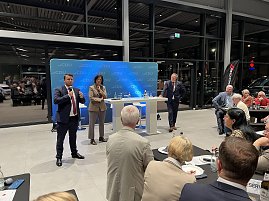 Drei Personen stehen vor einem CDU-Hintergrund auf einer Bühne im Autohaus. Eine Frau und zwei Männer sprechen in Mikrofone, während das Publikum aufmerksam zuhört. Auf den Tischen stehen Wasserflaschen und Gläser.