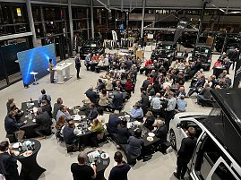 Blick von oben auf die Veranstaltung: zahlreiche Tische mit Gästen, die dem CDU-Wirtschaftsdialog folgen. Die Bühne steht links mit zwei Rednern, im Hintergrund mehrere Autos und Firmenbanner von Jeep und Dodge.