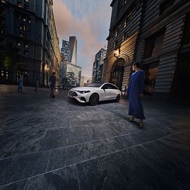 Das Bild zeigt einen weißen Mercedes-Benz CLA Shooting Brake, der auf einer breiten, nahezu leeren Stadtstraße fährt. Moderne Gebäude umgeben die Szene, das Licht wirkt weich und frühmorgendlich. Der Wagen ist seitlich zu sehen und vermittelt Dynamik sowie Eleganz.