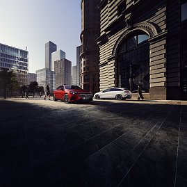 Das Bild zeigt einen weißen Mercedes-Benz CLA Shooting Brake, der auf einer breiten, nahezu leeren Stadtstraße fährt. Moderne Gebäude umgeben die Szene, das Licht wirkt weich und frühmorgendlich. Der Wagen ist seitlich zu sehen und vermittelt Dynamik sowie Eleganz.