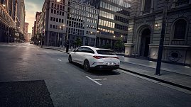 Das Bild zeigt einen weißen Mercedes-Benz CLA Shooting Brake, der auf einer breiten, nahezu leeren Stadtstraße fährt. Moderne Gebäude umgeben die Szene, das Licht wirkt weich und frühmorgendlich. Der Wagen ist seitlich zu sehen und vermittelt Dynamik sowie Eleganz.
