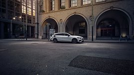 Das Bild zeigt einen weißen Mercedes-Benz CLA Shooting Brake, der auf einer breiten, nahezu leeren Stadtstraße fährt. Moderne Gebäude umgeben die Szene, das Licht wirkt weich und frühmorgendlich. Der Wagen ist seitlich zu sehen und vermittelt Dynamik sowie Eleganz.