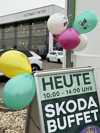 Werbeschild mit Text Heute 10:00–14:00 Uhr Skoda Buffet, dekoriert mit bunten Luftballons vor einem Autohaus.