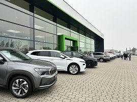 Reihe geparkter Autos vor einem modernen Autohaus mit großer Glasfront und grünem Eingang, Menschen gehen über den Parkplatz.