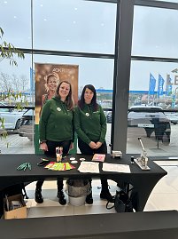 Zwei Frauen in grünen Hoodies stehen an einem Infotisch mit Stiften und Flyern, dahinter ein AOK-Plus-Banner.