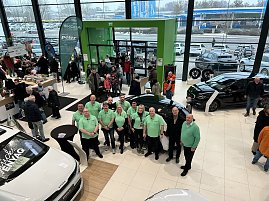 Mitarbeitergruppe in grünen Poloshirts steht im Autohaus zwischen ausgestellten Skoda-Fahrzeugen, im Hintergrund viele Besucher am Eingang.