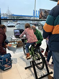 Kind sitzt auf einem Fahrradtrainer im Autohaus, daneben Erwachsene. Ein Laptop zeigt eine Strecke auf dem Bildschirm.