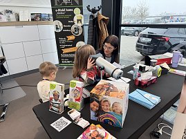 Eine Frau macht einen Sehtest mit einem kleinen Mädchen. Daneben sitzt ein kleiner Junge. Auf dem Tisch davor liegen Flyer, eine Box und Material.