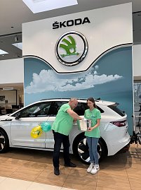 Mann und junge Frau in grünen Shirts stehen vor einem weißen Škoda im Autohaus. Die junge Frau hält einen silbernen Behälter, der Mann greift hinein und zieht einen Umschlag.