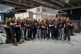 Gruppe von Mitarbeitenden posiert l&auml;chelnd mit Daumen hoch an einem Messestand in einer Messehalle.