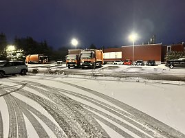 Winterliche Szene auf einem Betriebshof bei Nacht mit mehreren orangefarbenen M&uuml;llfahrzeugen und verschneitem Parkplatz.