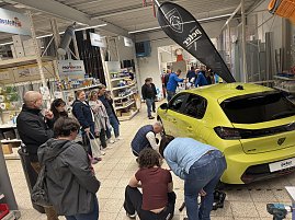 Gelber Peugeot im Baumarkt; viele interessierte Baumarktbesucher stehen rund herum und sehen beim R&auml;derwechsel zu.