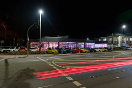 Au&szlig;enaufnahme bei Nacht: Ein modern verglastes Autohaus ist festlich beleuchtet, innen findet eine Veranstaltung statt. Davor stehen mehrere Fahrzeuge in einer Reihe.