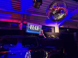 Stimmungsvolle Club-Inszenierung: Eine DJane performt hinter dem Pult, w&auml;hrend eine gro&szlig;e Discokugel das Licht in den Raum reflektiert.