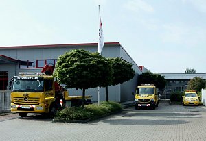 Drei gelbe ADAC-Fahrzeuge des Autohaus Peter, darunter ein Abschlepp-LKW, ein Transporter und ein PKW, stehen vor einem grauen Geb&auml;ude mit roten Fensterrahmen. Davor stehen zwei B&auml;ume und ein Fahnenmast mit Fahne. Der Himmel ist leicht bew&ouml;lkt.