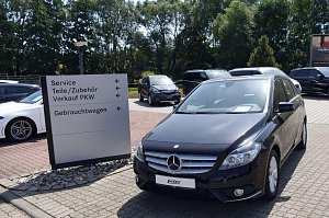 Parkplatz der Mercedes-Benz-Niederlassung in Duderstadt mit einem schwarzen Mercedes-Benz im Vordergrund. Daneben ein wei&szlig;es Auto und ein Wegweiser mit Beschriftungen wie Service, Teile/Zubeh&ouml;r, Verkauf PKW und Gebrauchtwagen. Im Hintergrund mehrere weitere Fahrzeuge und B&auml;ume.