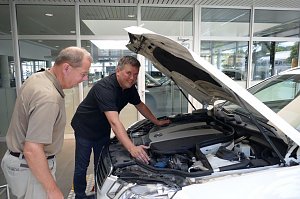 Zwei Männer stehen vor einem weißen Auto mit geöffneter Motorhaube. Einer der Männer trägt ein schwarzes Polohemd und zeigt dem anderen den Motorraum. Beide wirken konzentriert und interessiert.